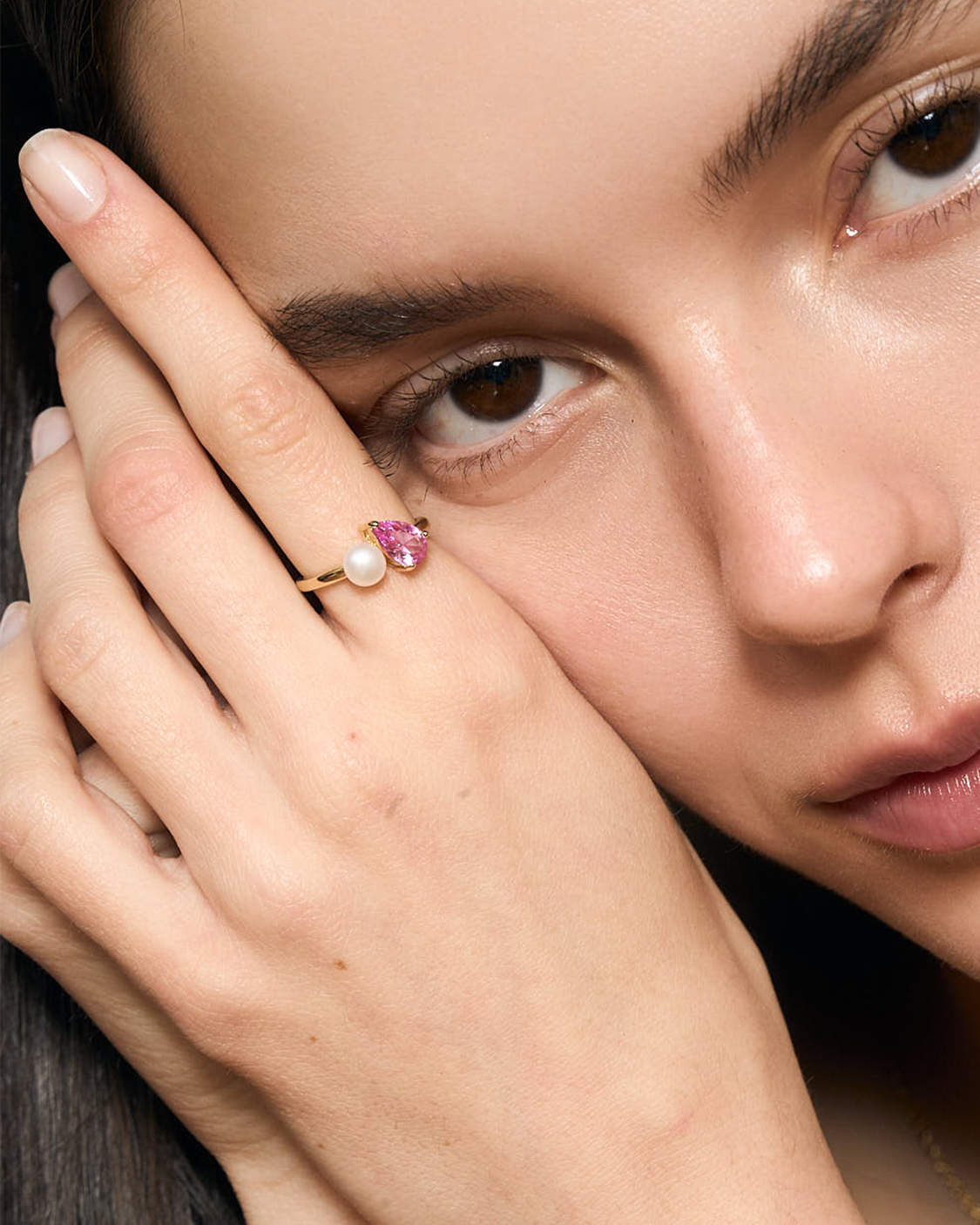 Close-up of a woman's face wearing a ring with a pink gemstone and pearl.