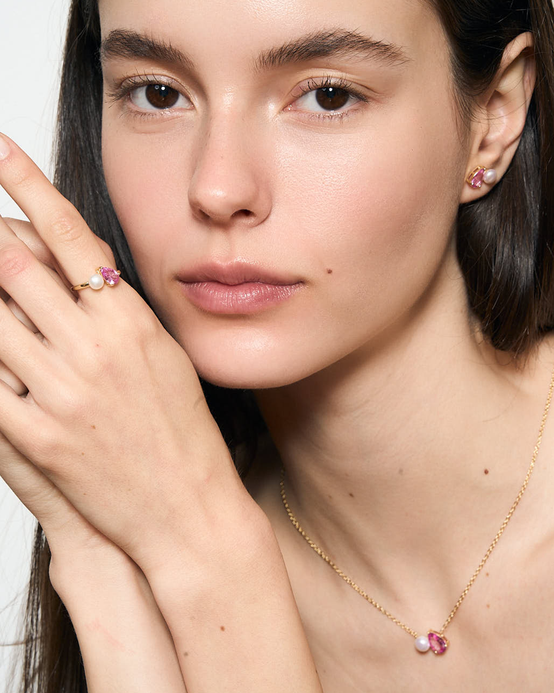 Close-up of a woman wearing a gold necklace with a pink pendant, a ring on her finger, and earrings.