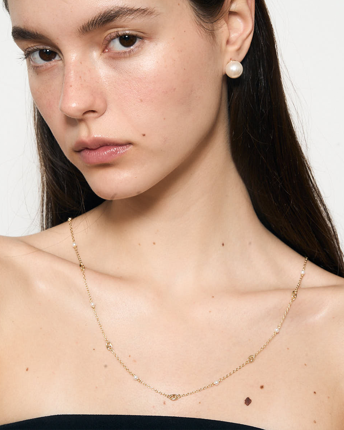Close-up of a woman wearing a gold necklace and pearl earrings against a white background
